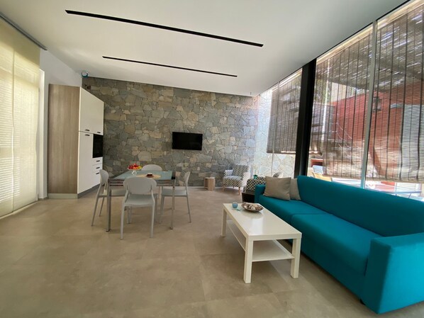 A modern living room with a stone wall, a teal sofa, a dining table with chairs, and a TV mounted on the wall.