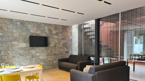 Una sala de estar moderna con una pared de piedra, un televisor de pantalla plana y un comedor con sillas amarillas y una mesa dispuesta para dos.