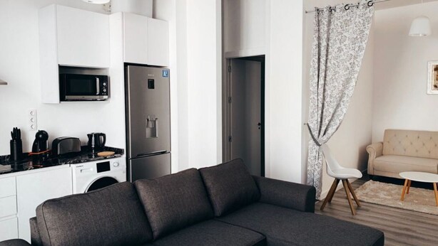 A modern living room with a grey sectional sofa, a black microwave, a stainless steel refrigerator, and a white ceiling light.