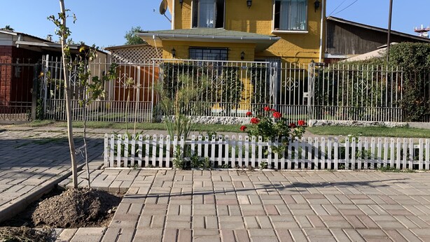 Una casa gialla a due piani con un recinto bianco a stecche, un vialetto asfaltato e un piccolo giardino con fiori rossi.