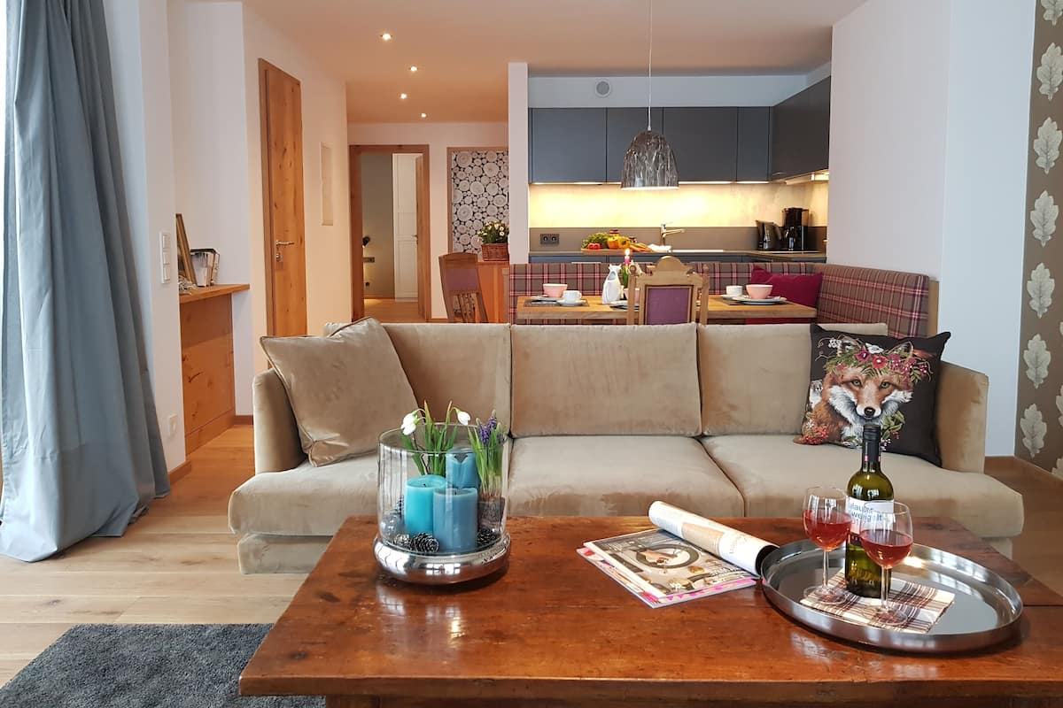 A living room with a sofa, a coffee table with drinks and a magazine, a kitchen in the background, and a dining area with a table set for two.