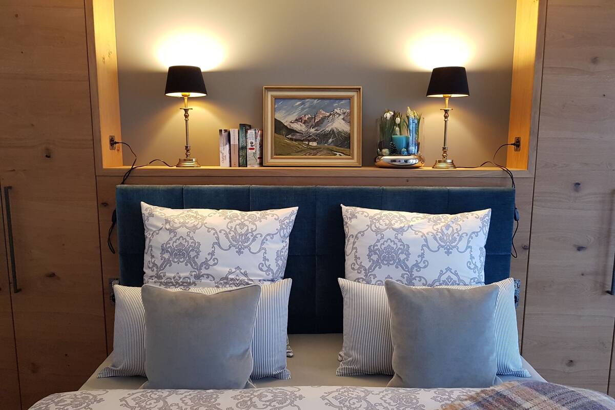 A bedroom with a large bed, two bedside lamps, a framed picture on the wall, and a shelf with books and decorative items.