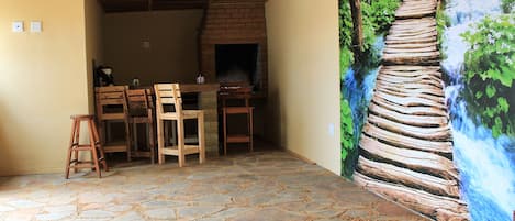 Un interior rústico con suelo de piedra, vigas de madera en el techo, chimenea de ladrillo y un mural que representa un puente de madera sobre una cascada.