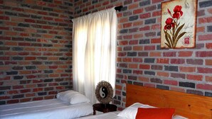 Un dormitorio con pared de ladrillo, una cama con ropa blanca y un cojín naranja, cabecera de madera, una mesita redonda con un objeto decorativo y una ventana con cortinas blancas.