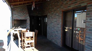 Un área de patio techada con pared de ladrillo, mobiliario de madera y puerta corrediza de vidrio.