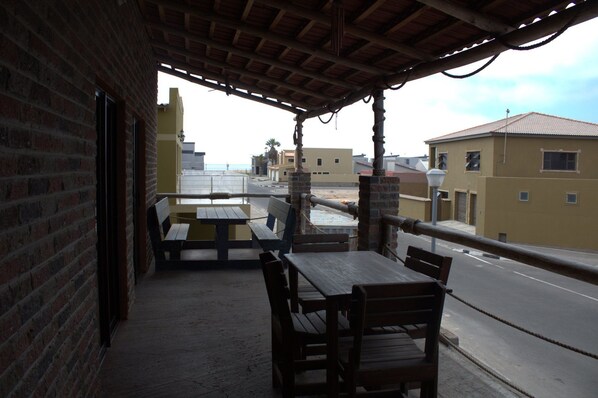 Un área de estar al aire libre con techo, con una mesa y bancos, con vistas a una calle con edificios y el mar.