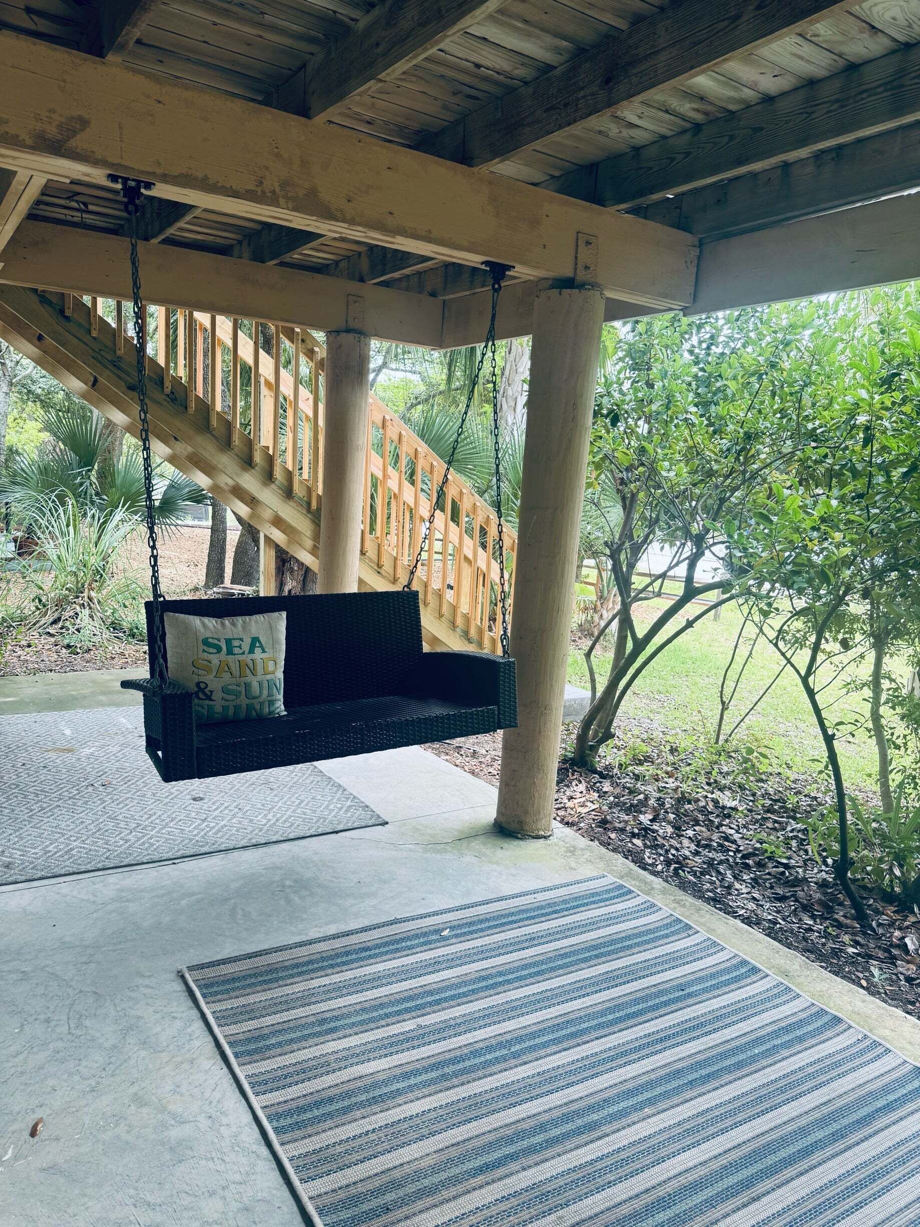 A covered outdoor area with a swing chair, a wooden staircase, and a view of greenery.