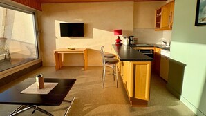 A compact kitchen with wooden cabinets, a dining area with a table and chairs, a built-in TV, and a large window.