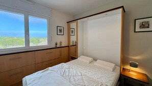 A bedroom with a bed, wooden wardrobe, a nightstand with a lamp, a large window, and framed pictures on the wall.