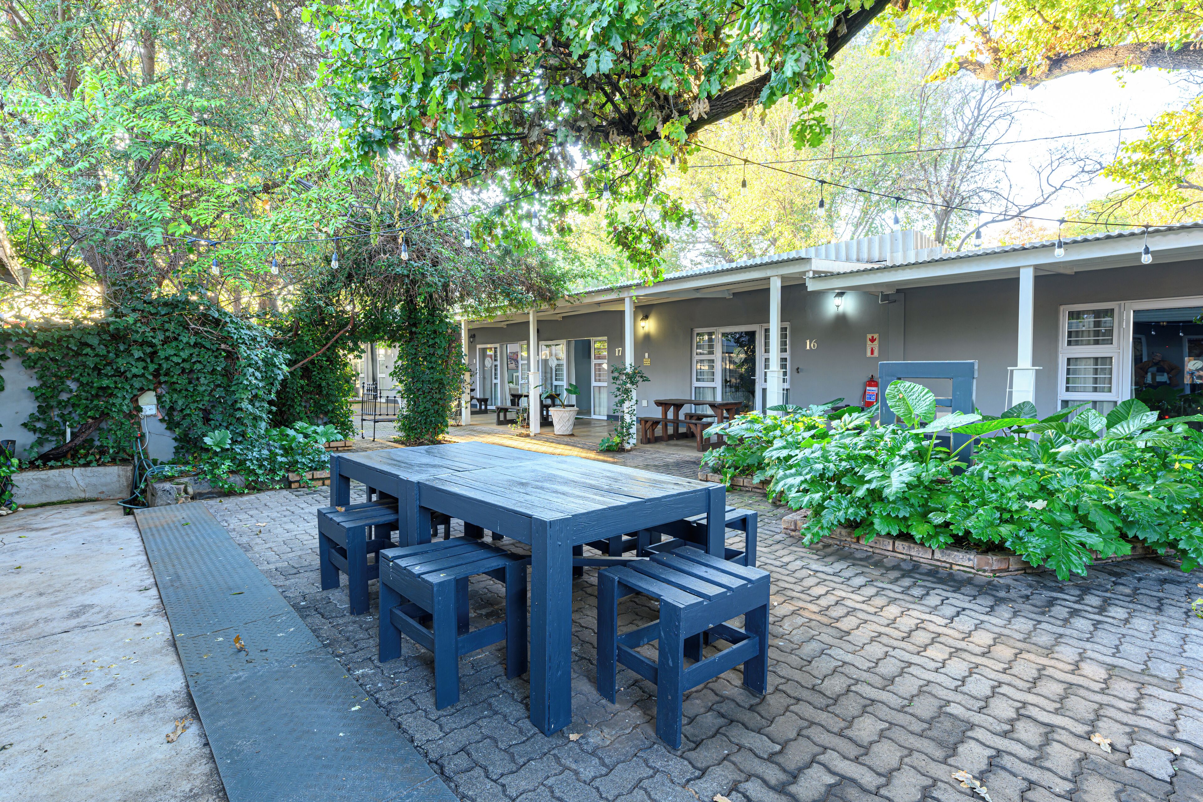 A patio area with a wooden table and benches, surrounded by greenery and a building with multiple windows and doors.