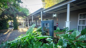 A covered outdoor area with a brick pathway, large green plants, and a building with multiple windows and a door. - Graceland Guest House (Potchefstroom)