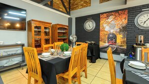 A dining area with wooden chairs and tables, a black brick wall with a clock, and a mural of a train. - Graceland Guest House (Potchefstroom)