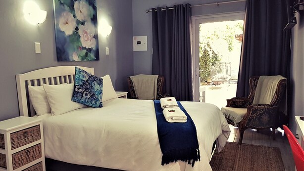 A bedroom with a bed, bedside tables, a chair, a painting, and a window with curtains. - Graceland Guest House (Potchefstroom)
