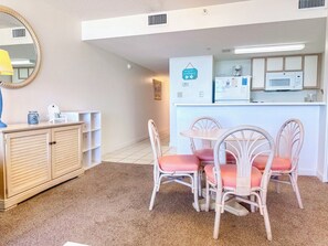 Un espacio habitable compacto con kitchenette, comedor con sillas de mimbre y una mesa pequeña, y un armario con estantes.