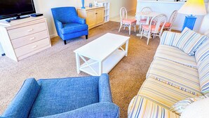 A living room with a TV, blue armchair, white sofa, and a dining area with chairs and a table.