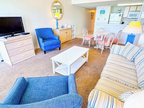 Una sala de estar con televisor, un sillón azul, un sofá a rayas, una mesa de centro blanca y una zona de cocina al fondo.