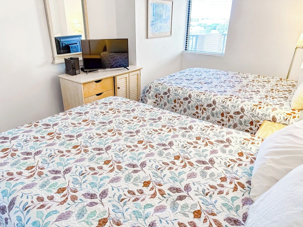 A bedroom with two beds, a desk with a computer, and a window with blinds.