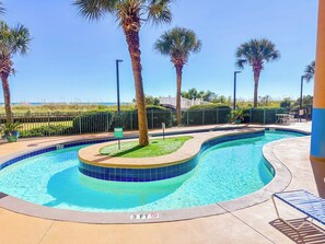 A swimming pool with a curved design, surrounded by palm trees and a fenced area. - Verandas 602 (North Myrtle Beach)