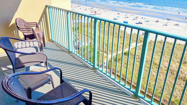 A balcony with two wicker chairs and a table, overlooking a beach with people and umbrellas.