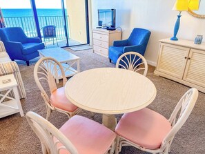 A room with a round table, four chairs, a television on a cabinet, and a view of the ocean. - Verandas 602 (North Myrtle Beach)