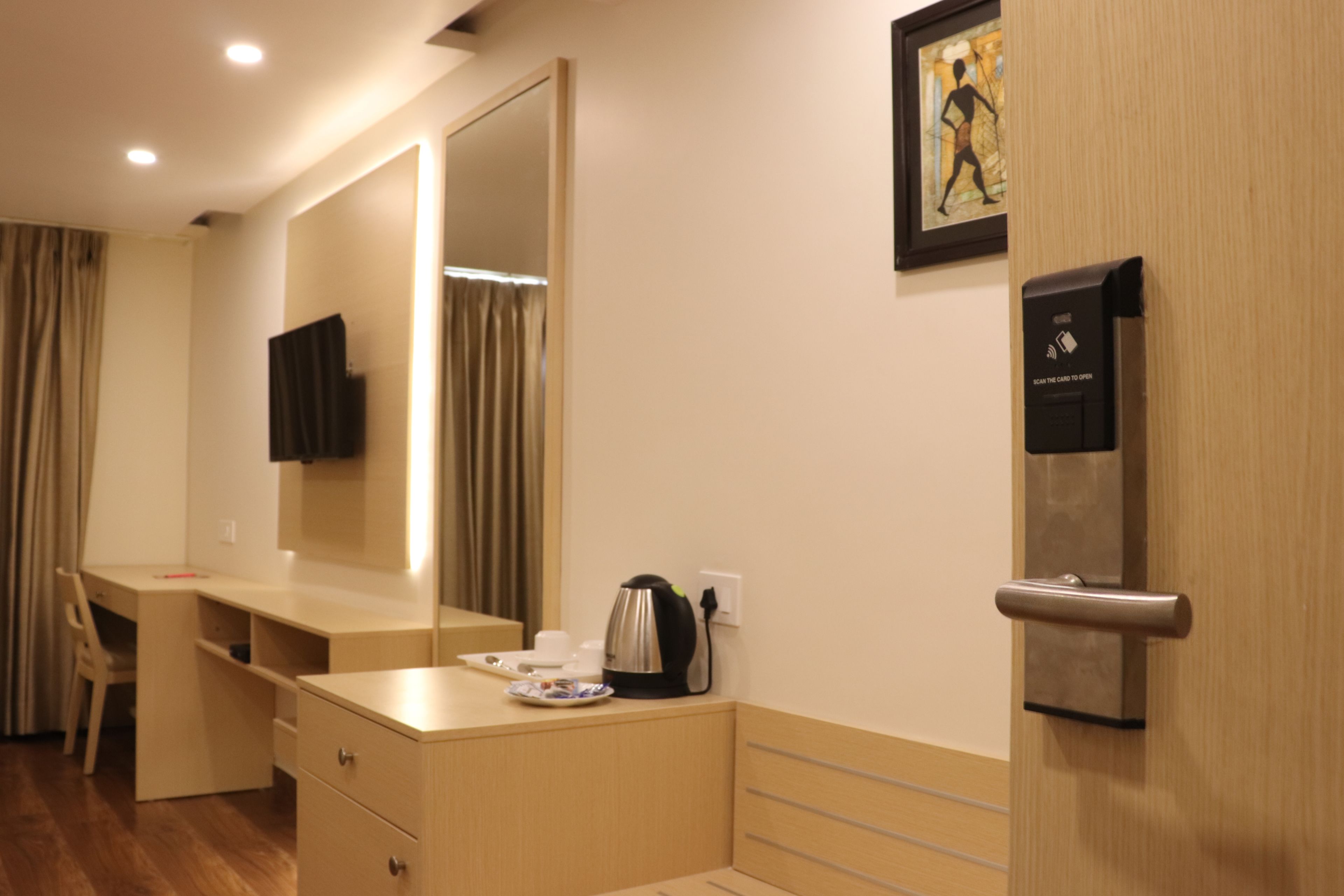A hotel room with a wooden desk, a large mirror, a coffee maker, a door with a card reader, and a framed picture on the wall.