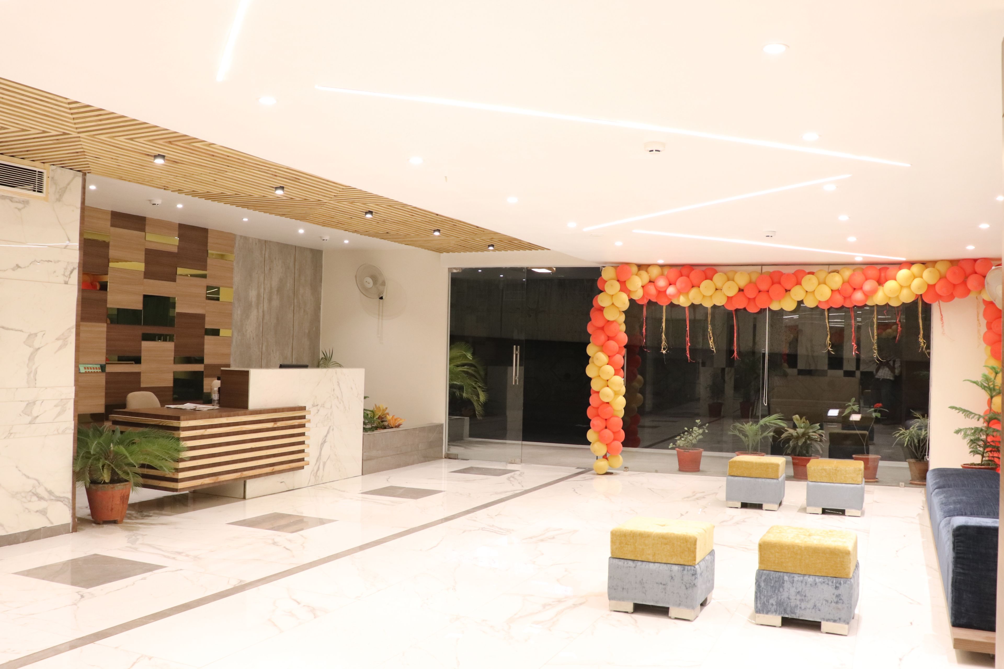 A modern reception area with a checkered wooden wall, a marble reception desk, and a colorful balloon arch. There are potted plants and a blue sofa in the space.