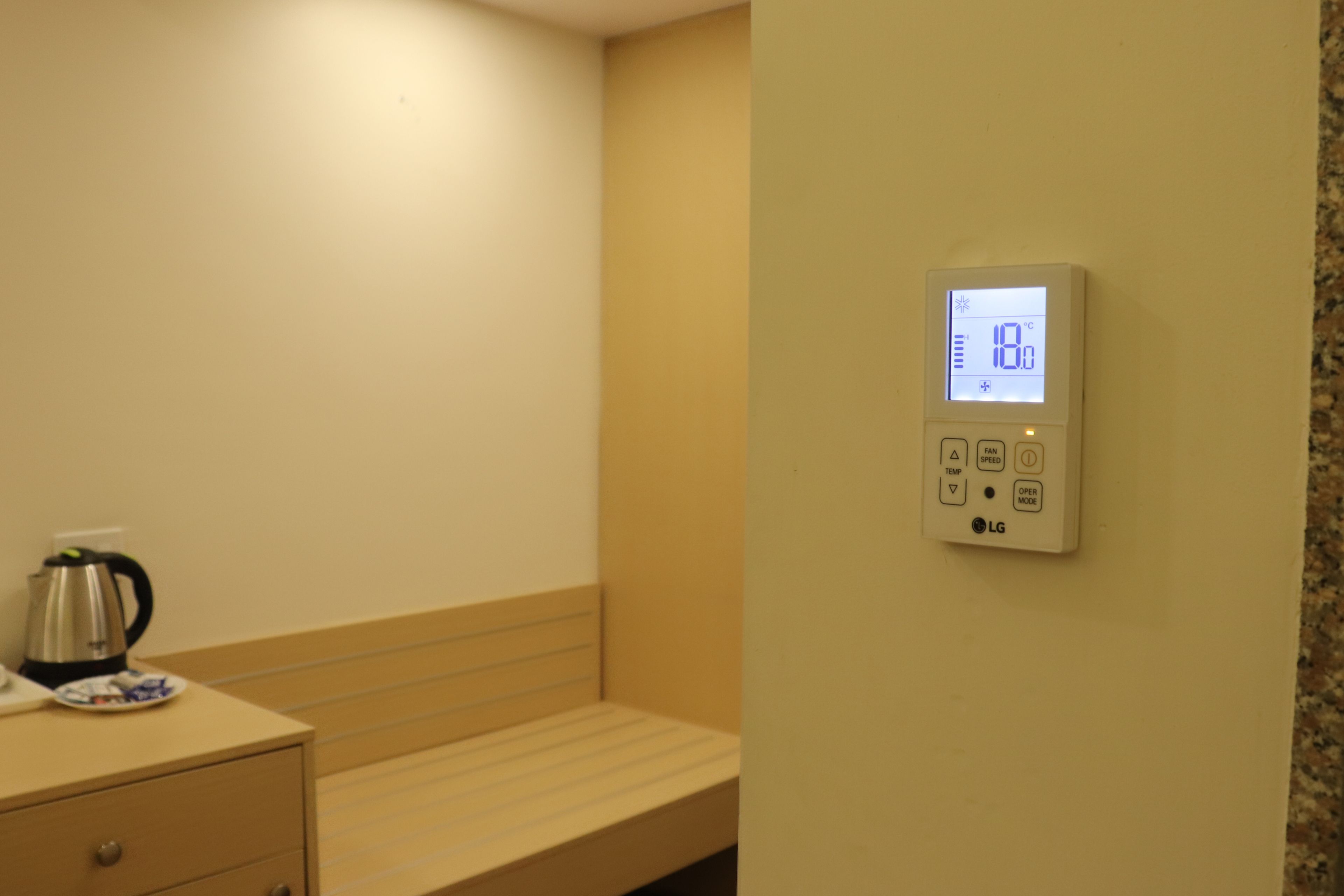 A hotel room with a bed, a bedside table with a kettle, and a thermostat on the wall.