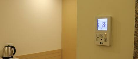 A hotel room with a bed, a bedside table with a kettle, and a thermostat on the wall.