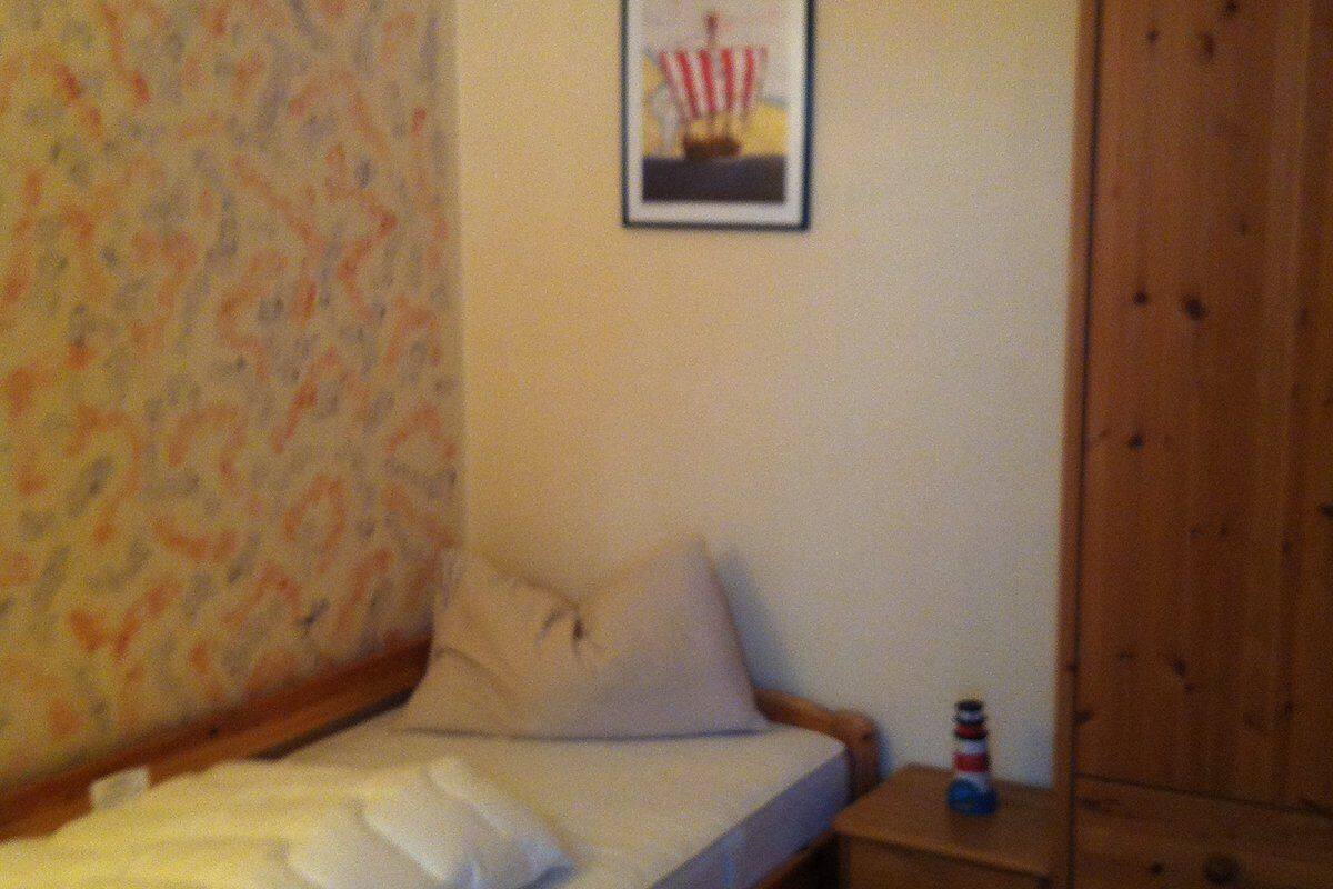 Una habitación pequeña y sencilla con una cama, una mesita de noche de madera y un cuadro enmarcado en la pared.