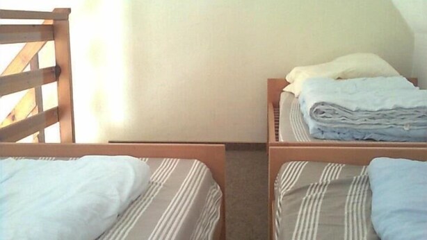 Un alojamiento pequeño y de una sola habitación con dos camas individuales, una silla de madera, un cuadro enmarcado en la pared y una ventana que proporciona luz natural.