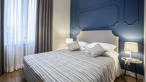 A neatly made bed with a striped bedspread, a bedside table with a lamp, and a view of a window with curtains. - Hotel Il Sole (Empoli)