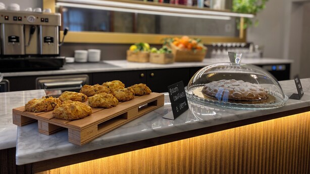 A bakery display with scones and a covered cake, a coffee machine, and shelves with various bottles in the background. - Hotel Il Sole (Empoli)