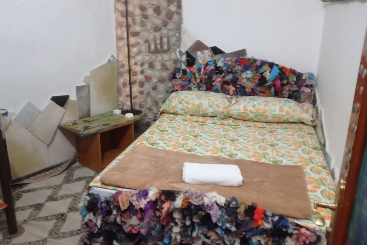 Une petite chambre à coucher, bien rangée, avec un lit, une table de chevet, un mur décoré d’un motif imitant la pierre et un rideau coloré.