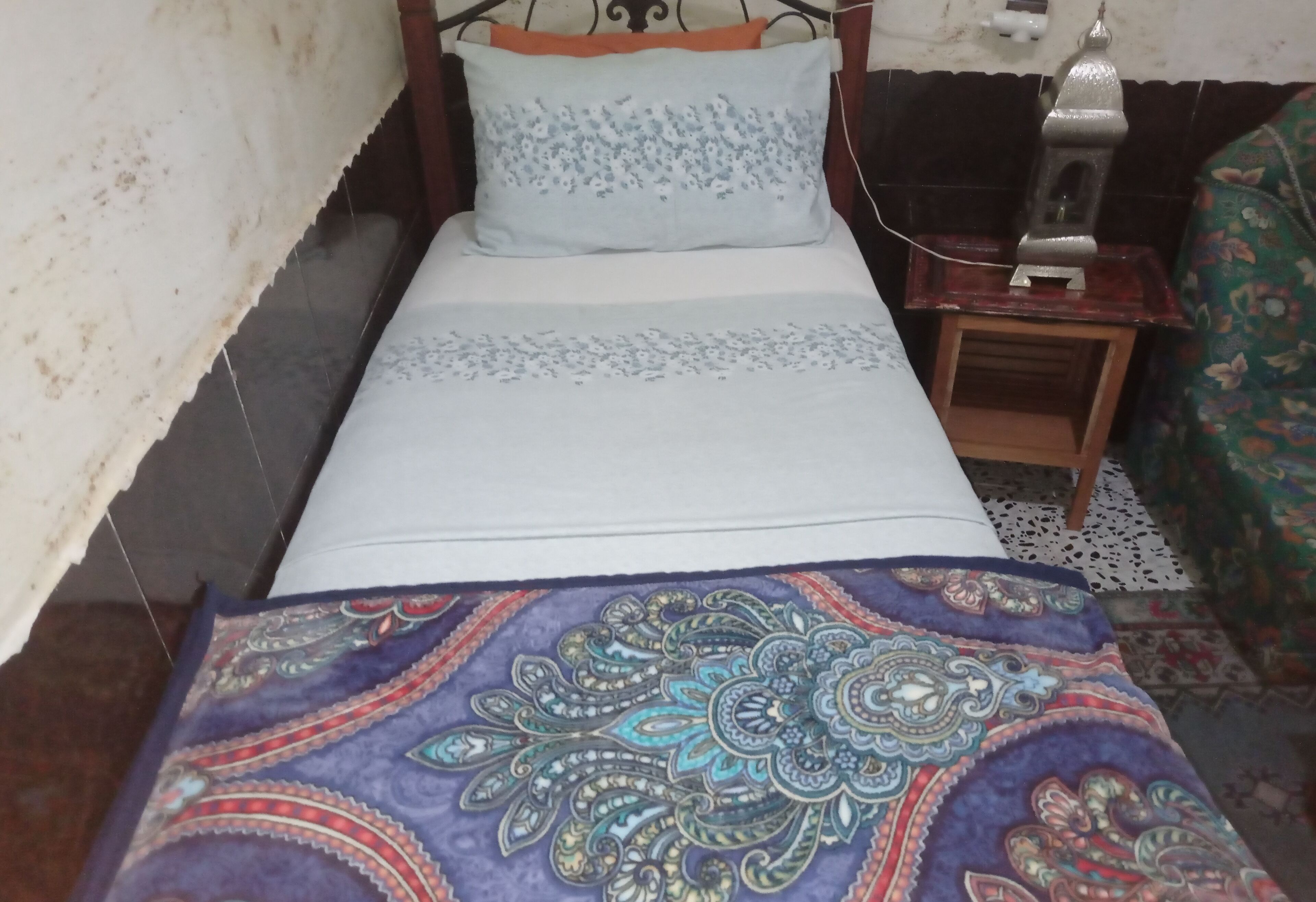 A bed with a patterned bedspread, a bedside table with a lamp, and a wall-mounted switch.