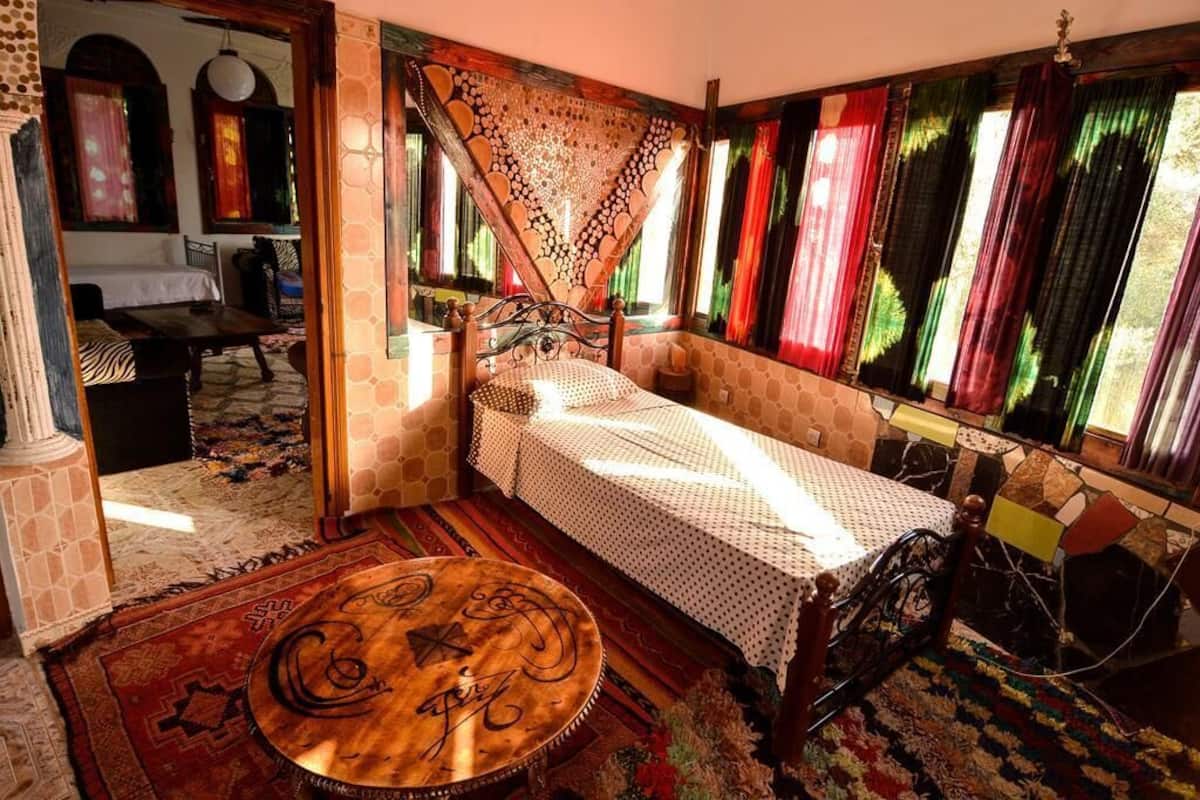 Une pièce chaleureuse avec un lit, une table ronde en bois et une fenêtre agrémentée de rideaux colorés.