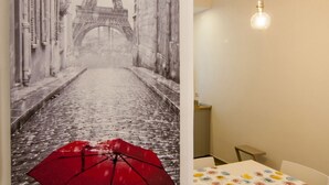 Une pièce ornée d’une grande fresque murale représentant la Tour Eiffel, d’une table recouverte d’une nappe à motifs et de deux luminaires suspendus.