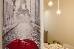 Une pièce ornée d’une grande fresque murale représentant la Tour Eiffel, d’une table recouverte d’une nappe à motifs et de deux luminaires suspendus.