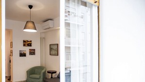 Une pièce avec un fauteuil vert, une petite table ronde, un luminaire suspendu et une fenêtre donnant sur un immeuble.