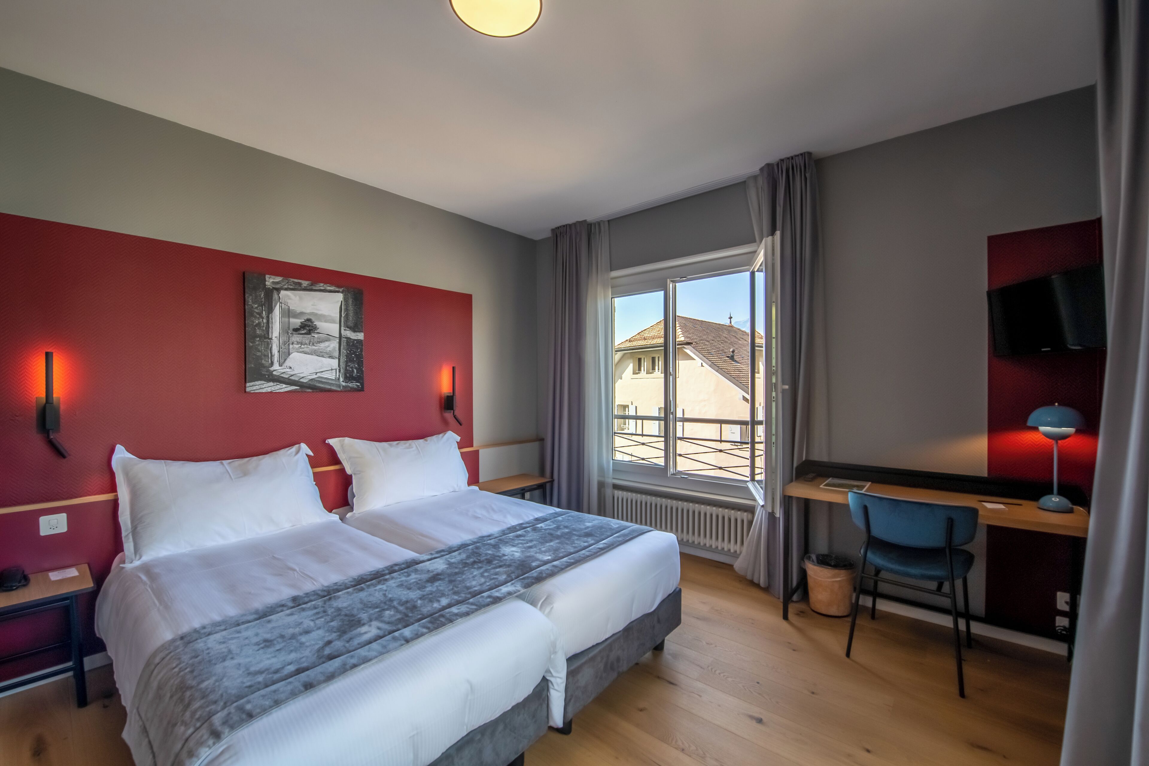 Une chambre d’hôtel équipée d’un lit, d’un bureau, d’une chaise, d’une télévision et d’une fenêtre donnant sur des immeubles.