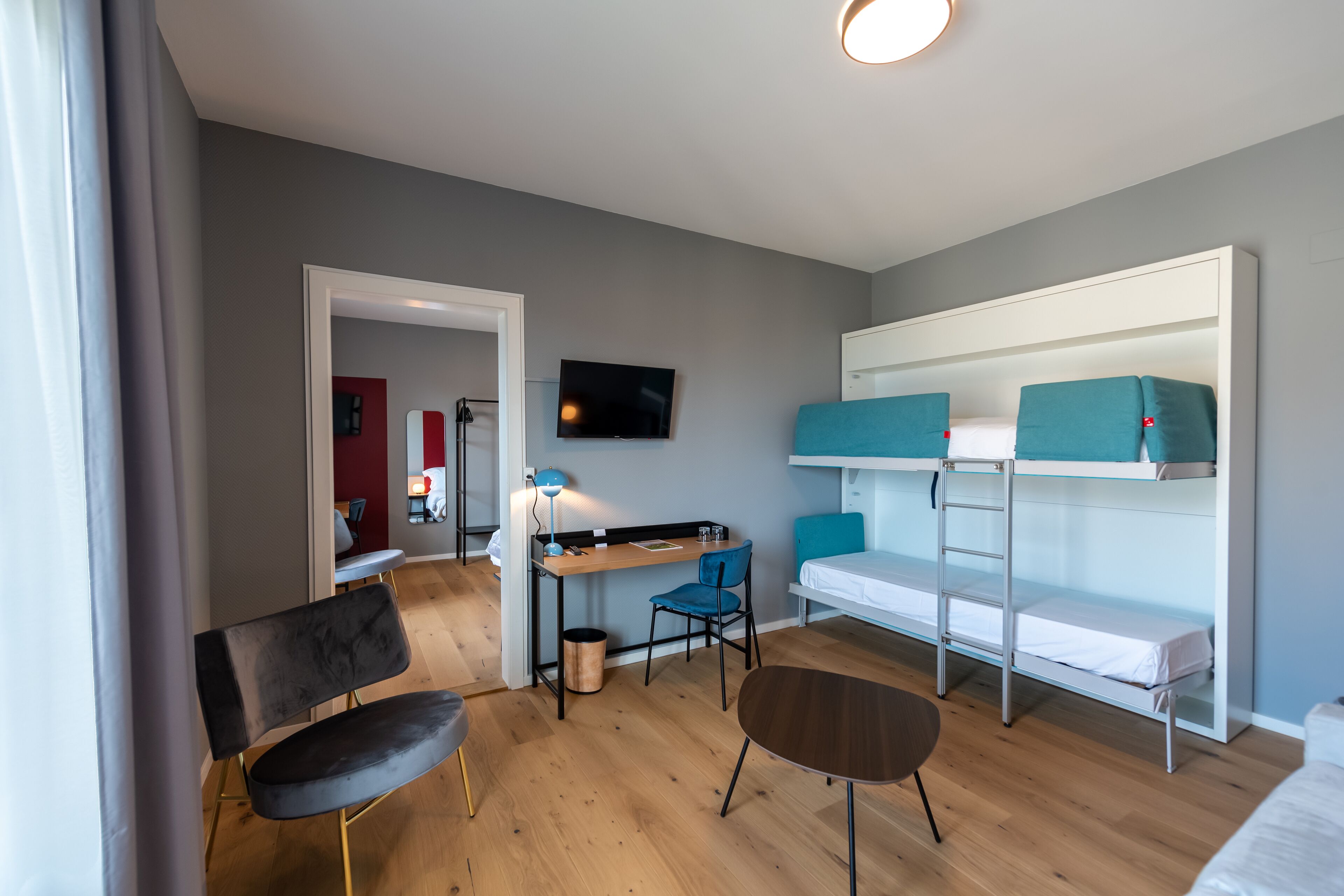 A modern, compact room with a bunk bed, a desk with a chair, a small table, and a TV mounted on the wall.