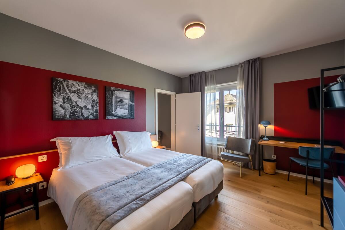 Una habitación de hotel moderna con una cama grande, un escritorio y una silla. Hay dos cuadros en la pared, una ventana con cortinas y un televisor sobre el escritorio.