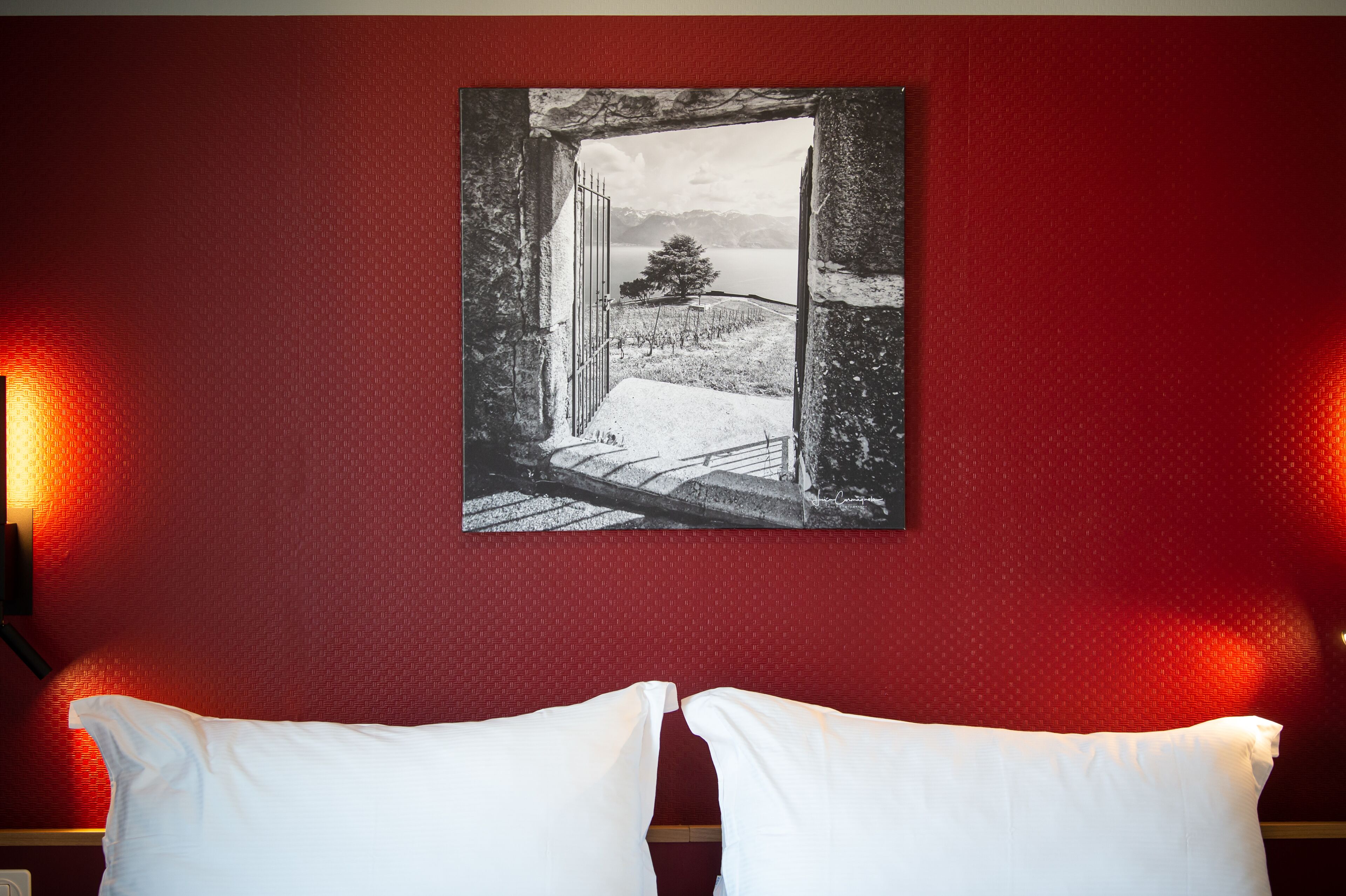 A room with a red textured wall, a framed black and white landscape painting depicting a snowy landscape with a tree, and two white pillows on a bed.