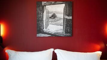 Una habitación con pared roja texturizada, un cuadro en blanco y negro con un paisaje nevado que muestra un árbol, y dos almohadas blancas sobre una cama.
