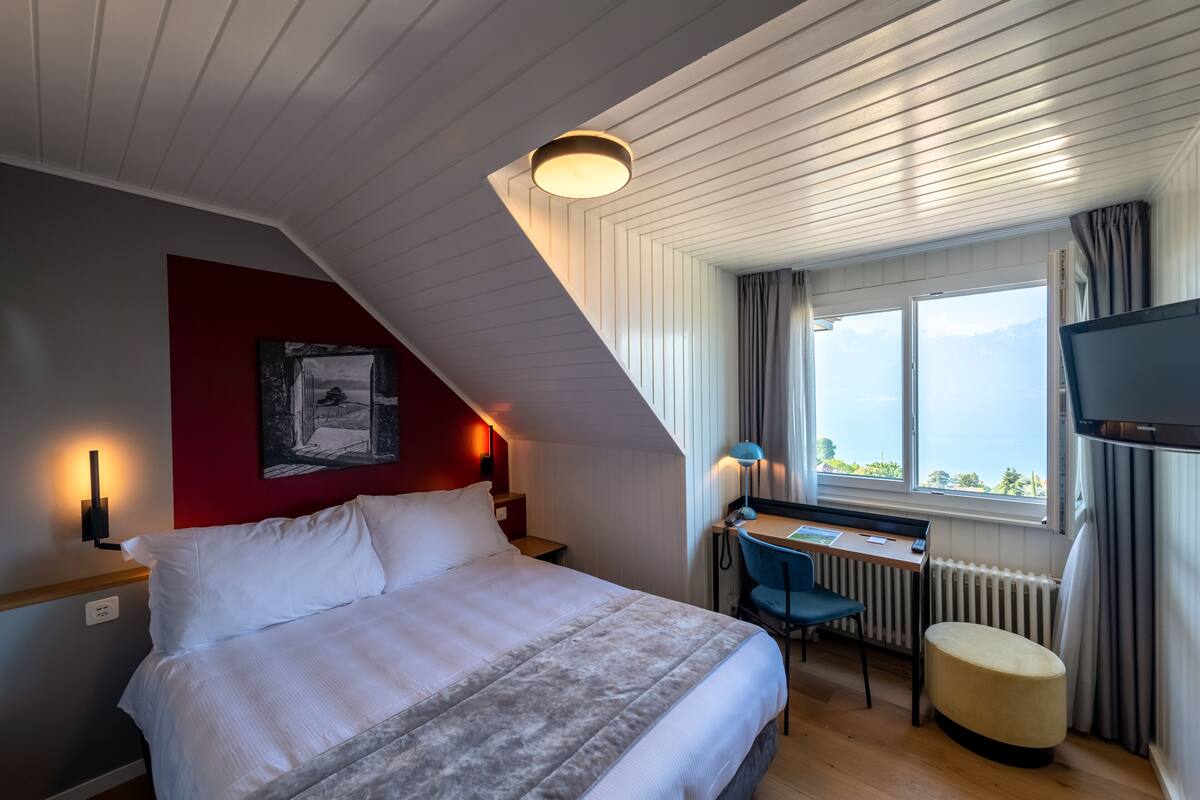 Una habitación de hotel moderna con una cama grande, un escritorio, una silla, un televisor y una ventana con vistas a la montaña.