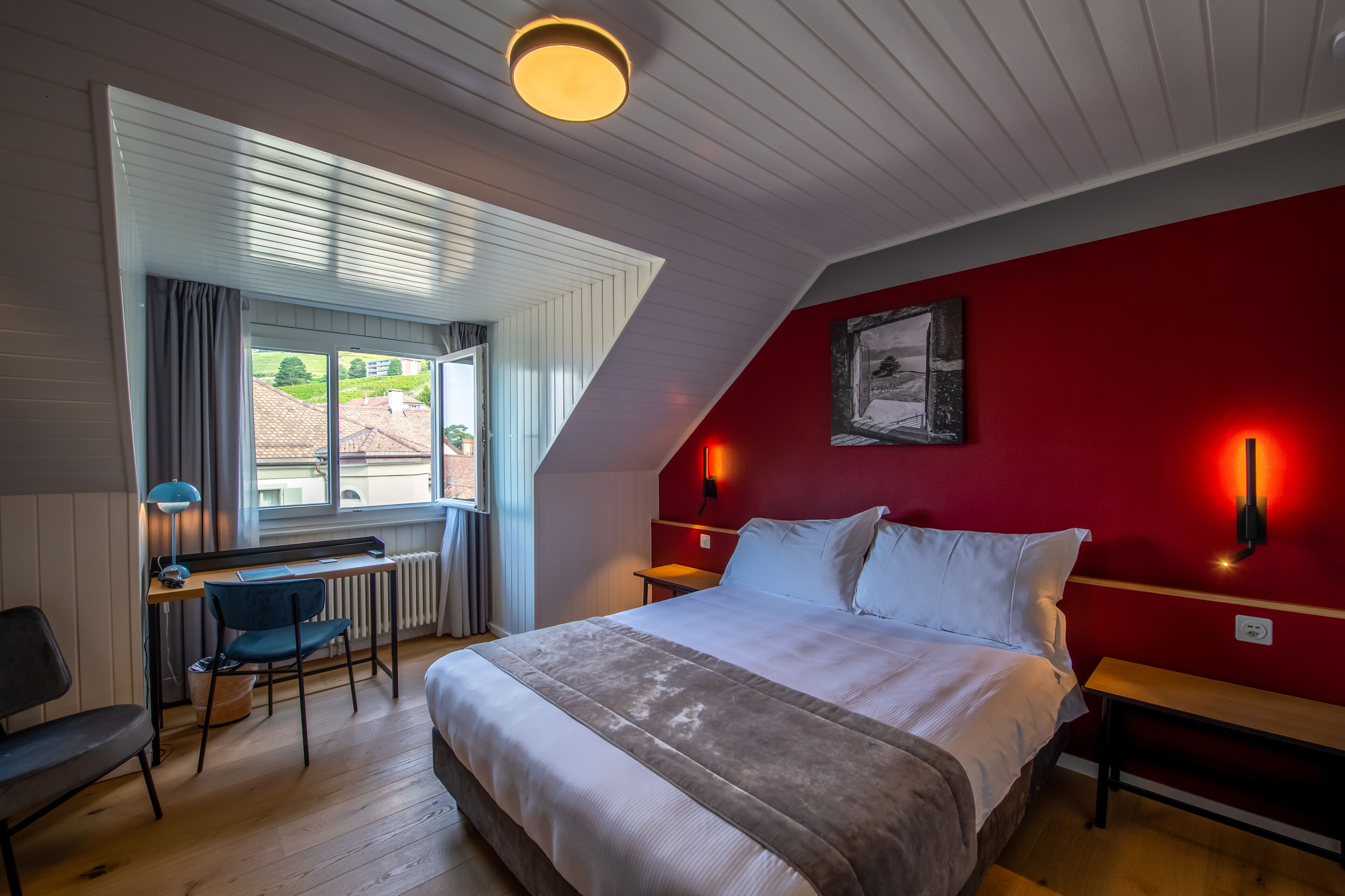 A bedroom with a bed, a desk, and a chair. There is a window with curtains, a red wall, and a picture on the wall.