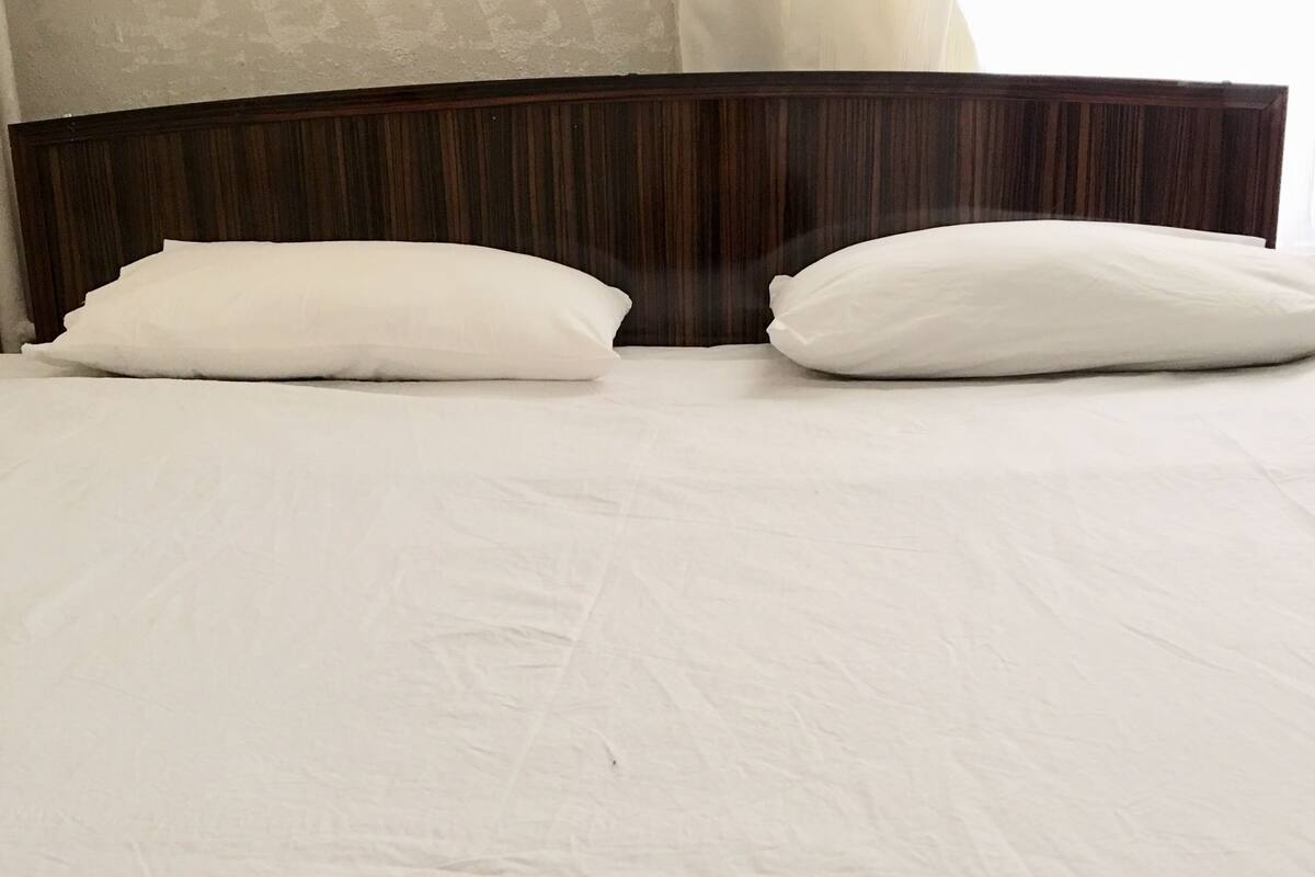 A neatly made bed with white linens and two pillows, a dark wooden headboard, and a light-colored wall with a subtle pattern.