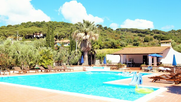 A swimming pool with a slide, surrounded by lounge chairs and umbrellas, set against a backdrop of a hillside with trees and a building. - Azienda Agricola Sapereta (Porto Azzurro)