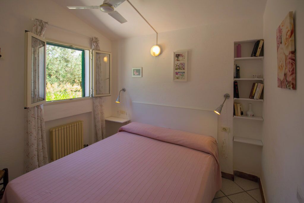 A bedroom with a bed, a window with curtains, a radiator, a wall-mounted shelf with books, and a ceiling fan.