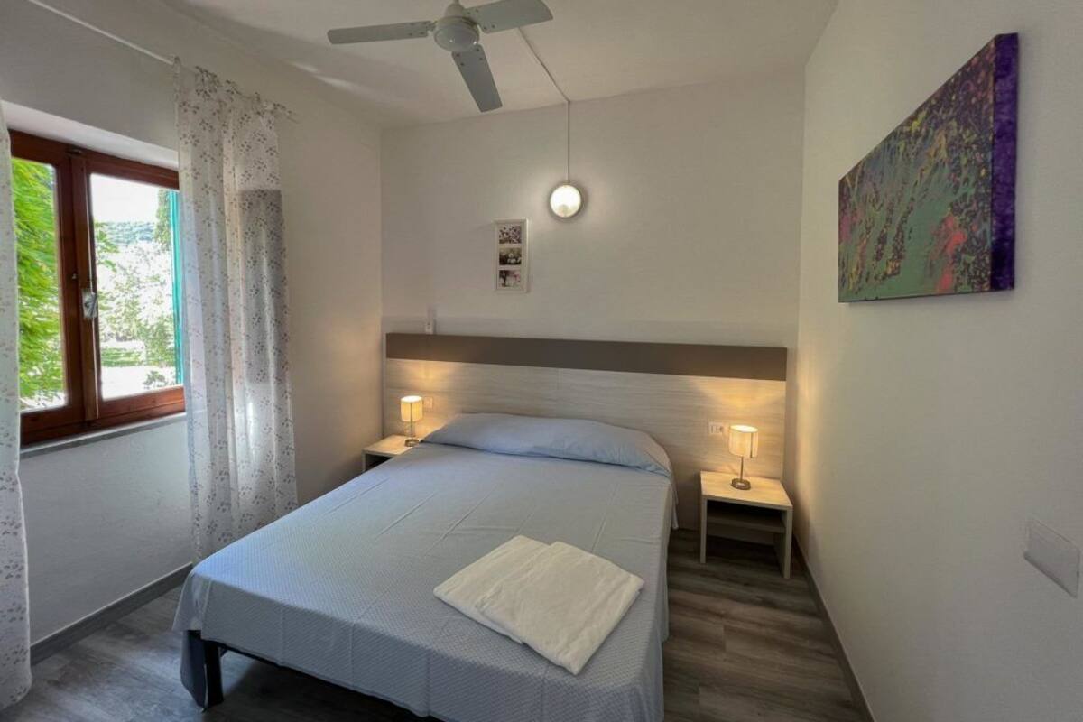 A bedroom with a bed, two bedside lamps, a ceiling fan, a window with curtains, and a painting on the wall.