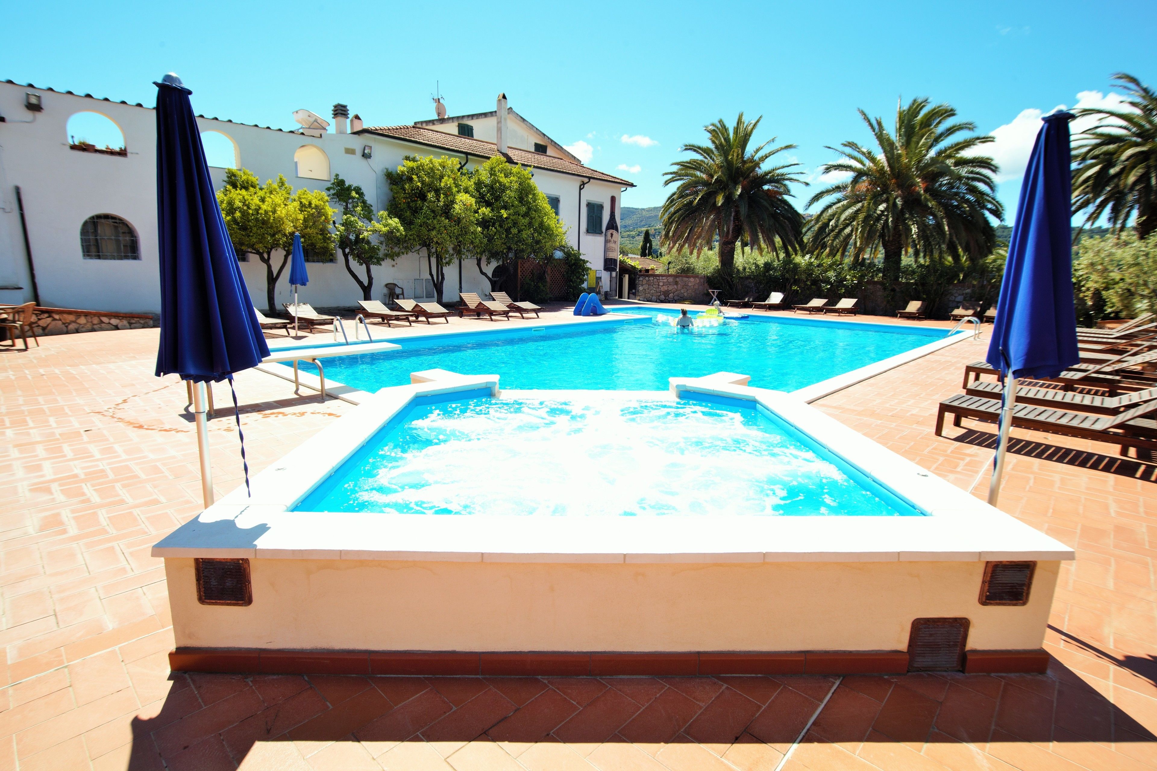 A rectangular swimming pool with a hot tub, surrounded by lounge chairs and umbrellas, set against a backdrop of a white building with arched windows and palm trees.